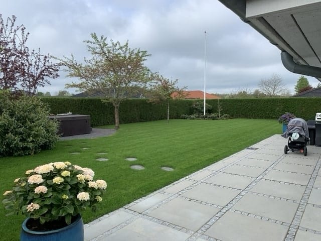 Udpluk af haven på en grund - et flot haveanlæg med fliser, planter og en grøn græsplæne.